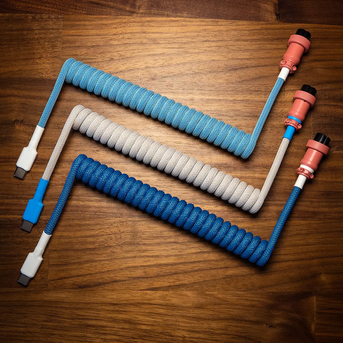 Three custom keyboard cables are displayed on a wooden surface, featuring coiled paracord textures. Each cable has detachable connectors—USB-C and aviator. The vibrant blue and gray colors are complemented by precision CNC metal housings and quality braiding.