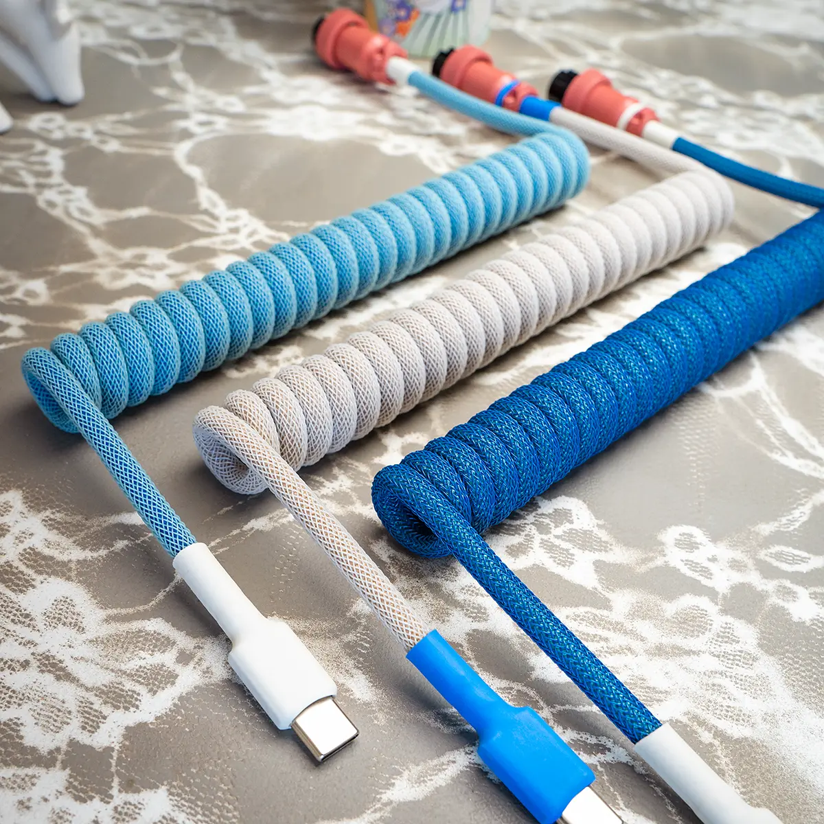 A collection of custom keyboard cables in various shades of blue and gray. Features include a coiled design, USB-C connectors, and textured paracord braiding.