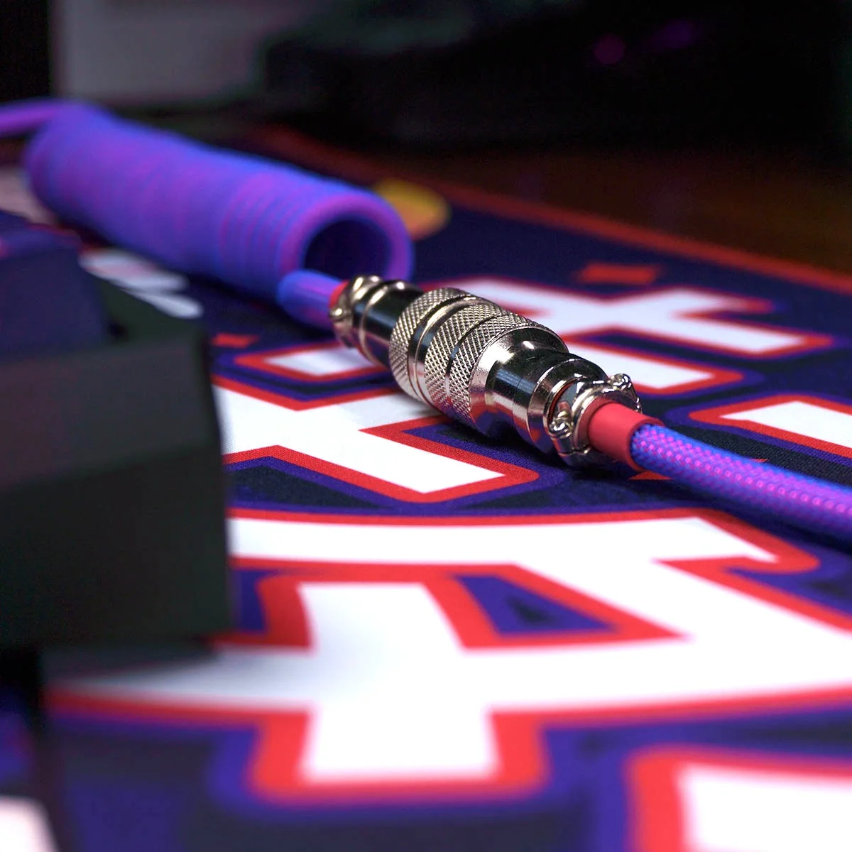 A close-up view of a laser-themed coiled purple cable with a GX16 silver 4-pin connector, lying on a colorful surface with bold text and graphics.