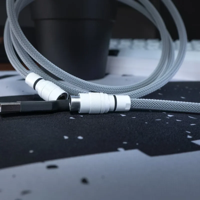 A coiled, black & white USB cable with yc8 connector white and sleek connectors rests on a black and gray patterned mouse pad, with a black cup and keyboard blurred in the background.