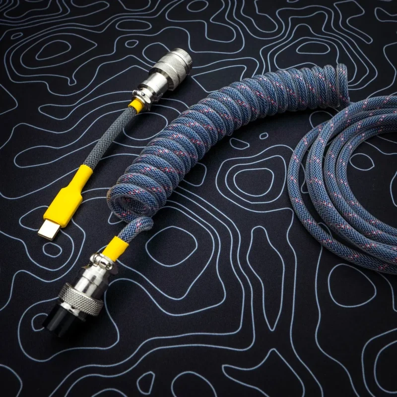 A dark blue coiled keyboard cable featuring a USB-C connector with a yellow accent, a detachable aviator plug, and quality braiding for durability.
