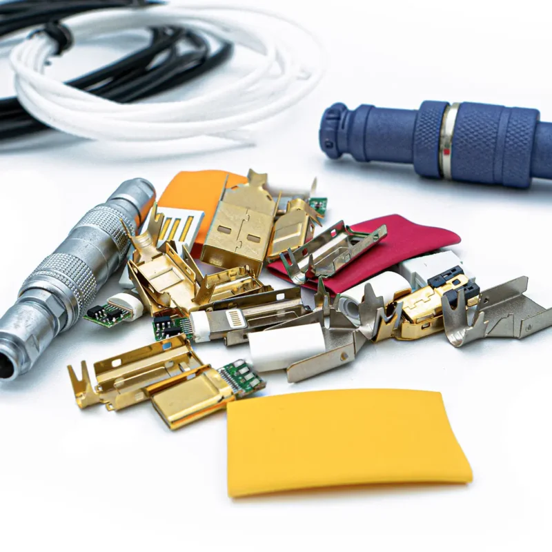 Assorted electronic and metal connector parts, various electronic components, and connectors in a plastic bag, including three colored wires, are arranged on a white surface.