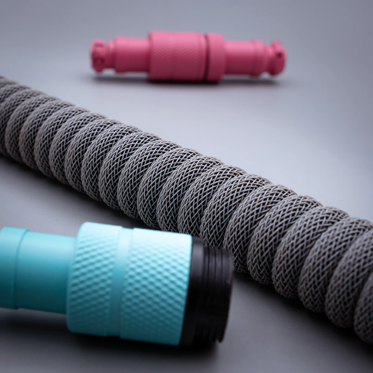 Custom keyboard cable featuring a textured gray coil, with turquoise and pink detachable connectors, ideal for versatile setups. Quality braiding ensures durability.