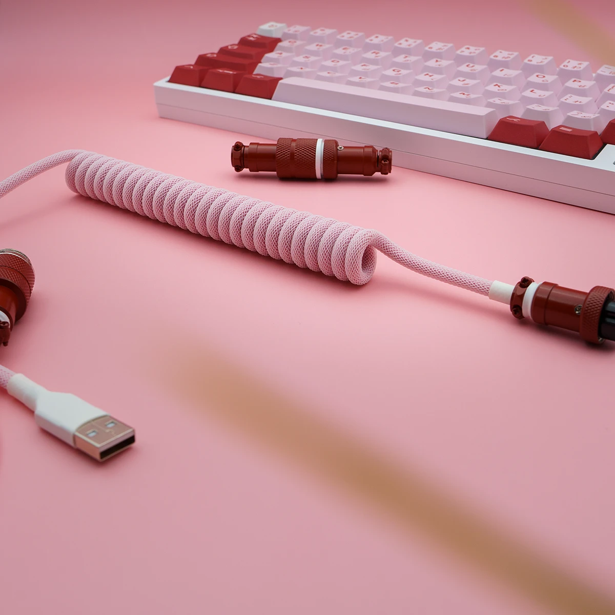 Coiled pink paracord keyboard cable with USB-C connector, detachable aviator plugs, and textured red metal housings, set against a pink background.