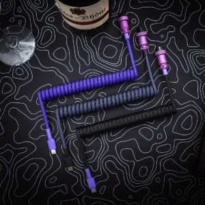 Three custom keyboard cables displayed on a textured background: one purple coiled, one black coiled, and a black straight cable. Each features detachable USB-C connectors and vibrant paracord.