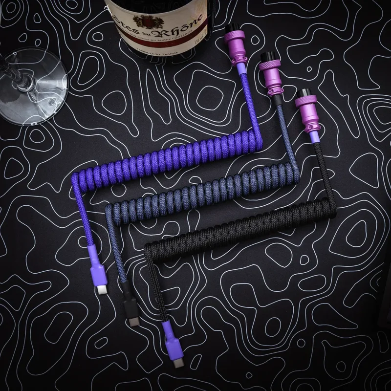 Three custom keyboard cables displayed on a textured background: one purple coiled, one black coiled, and a black straight cable. Each features detachable USB-C connectors and vibrant paracord.
