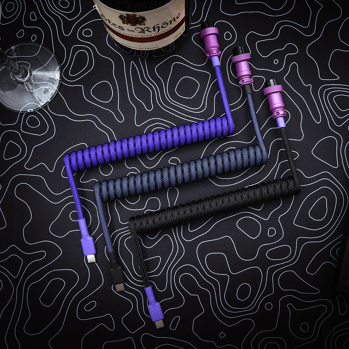 Three custom keyboard cables displayed on a textured background: one purple coiled, one black coiled, and a black straight cable. Each features detachable USB-C connectors and vibrant paracord.