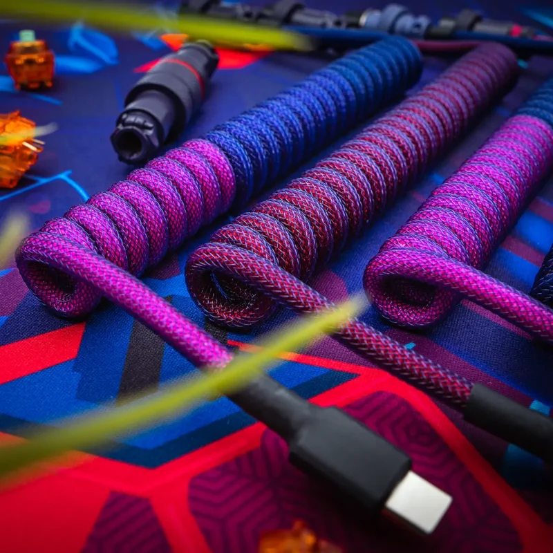 Three coiled custom keyboard cables showcase vibrant color schemes in purple and blue paracord. Features include USB-C connectors and high-quality braiding.