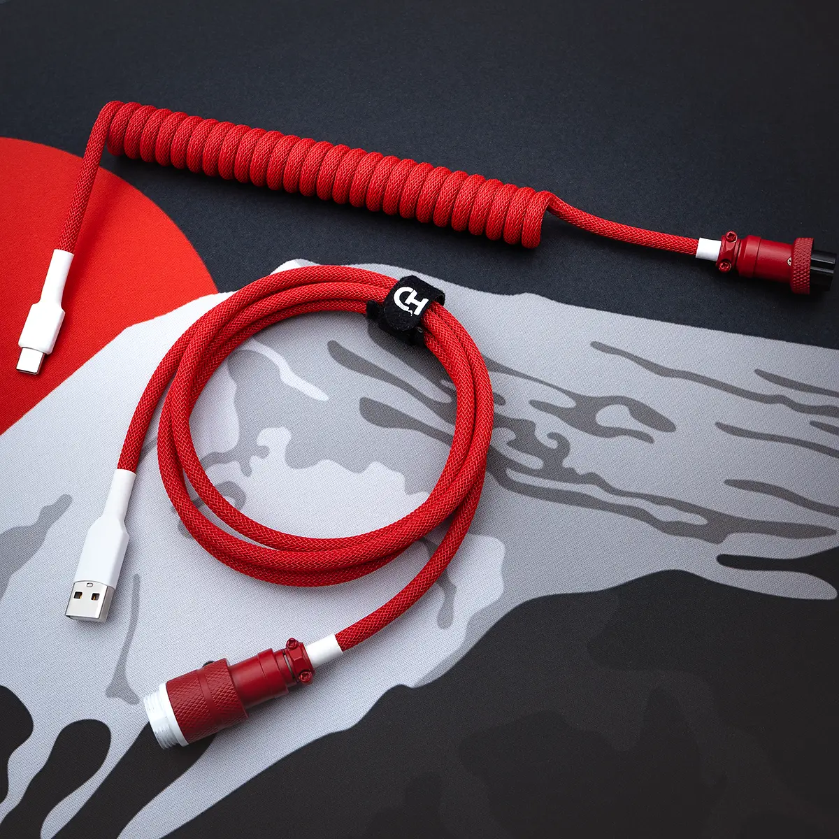 A vibrant red GMK Jamon cable features a coiled design with a USB-C connector and a detachable aviator plug, showcasing premium paracord braiding and heatshrink.