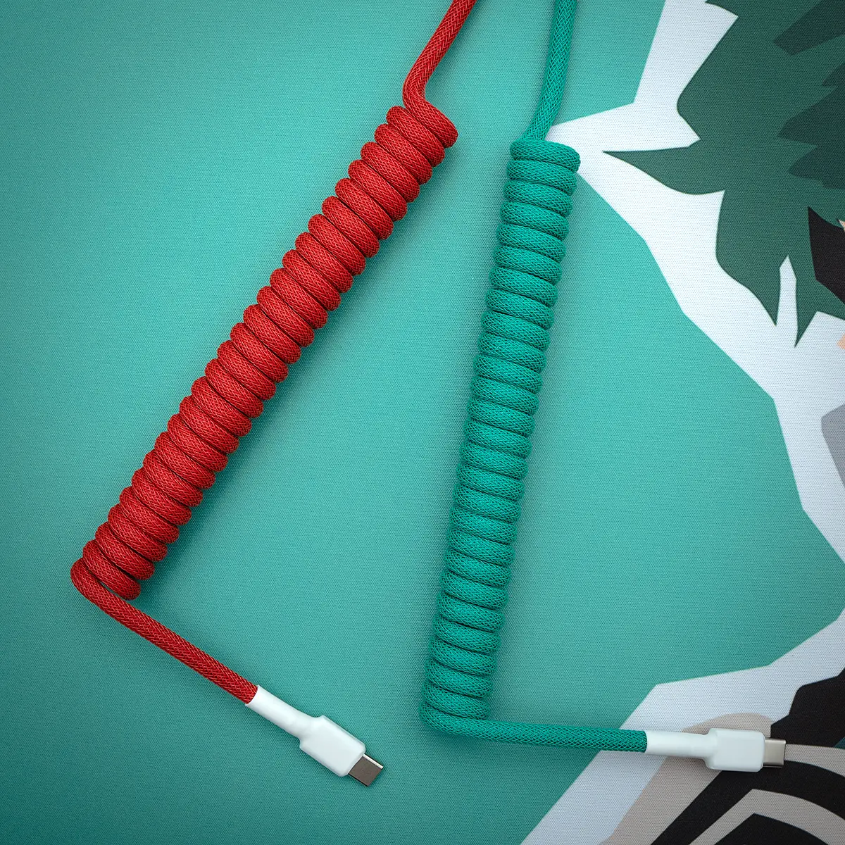 Two custom keyboard cables in red and green paracord with coiled sections and USB-C connectors, showcasing quality braiding and finish details.
